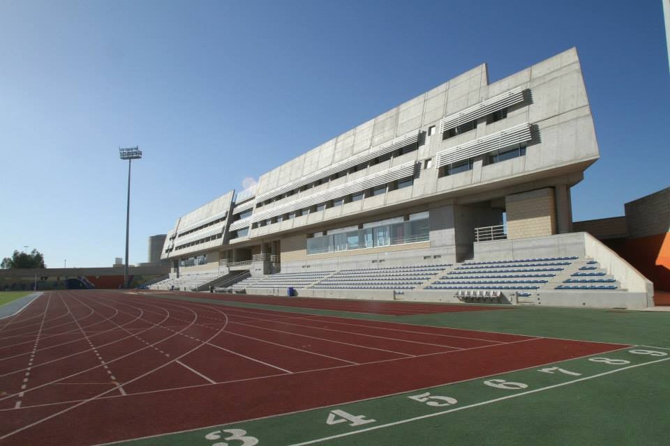 Modern athletics stadium with red running track and large grandstand under clear blue sky