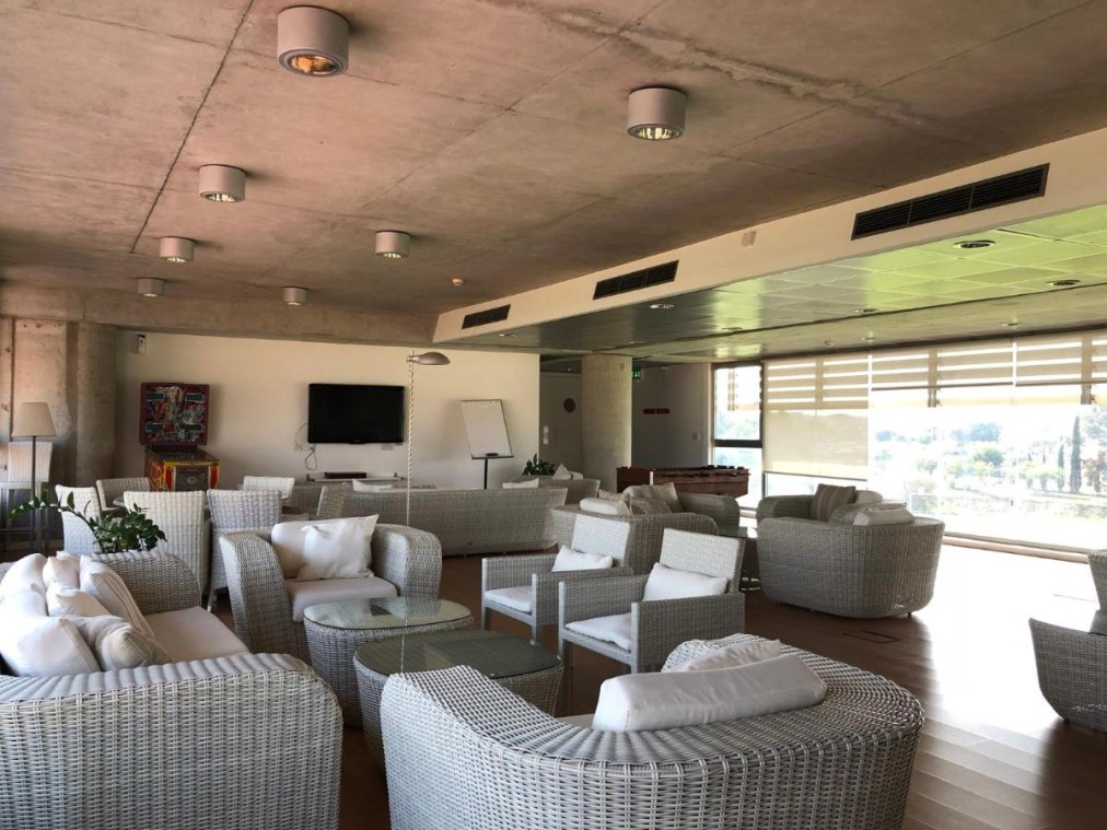 Lounge area with wicker furniture, white cushions, and large windows overlooking sports facilities