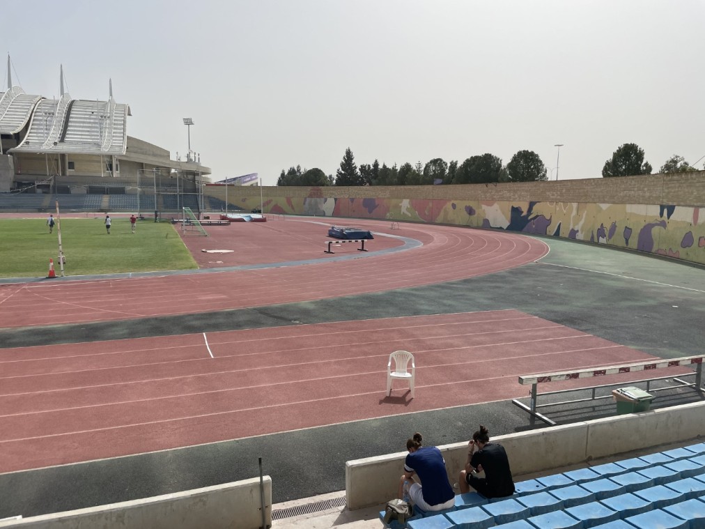 Athletics facility with curved red track, colorful wall mural, and athletes resting on blue stands