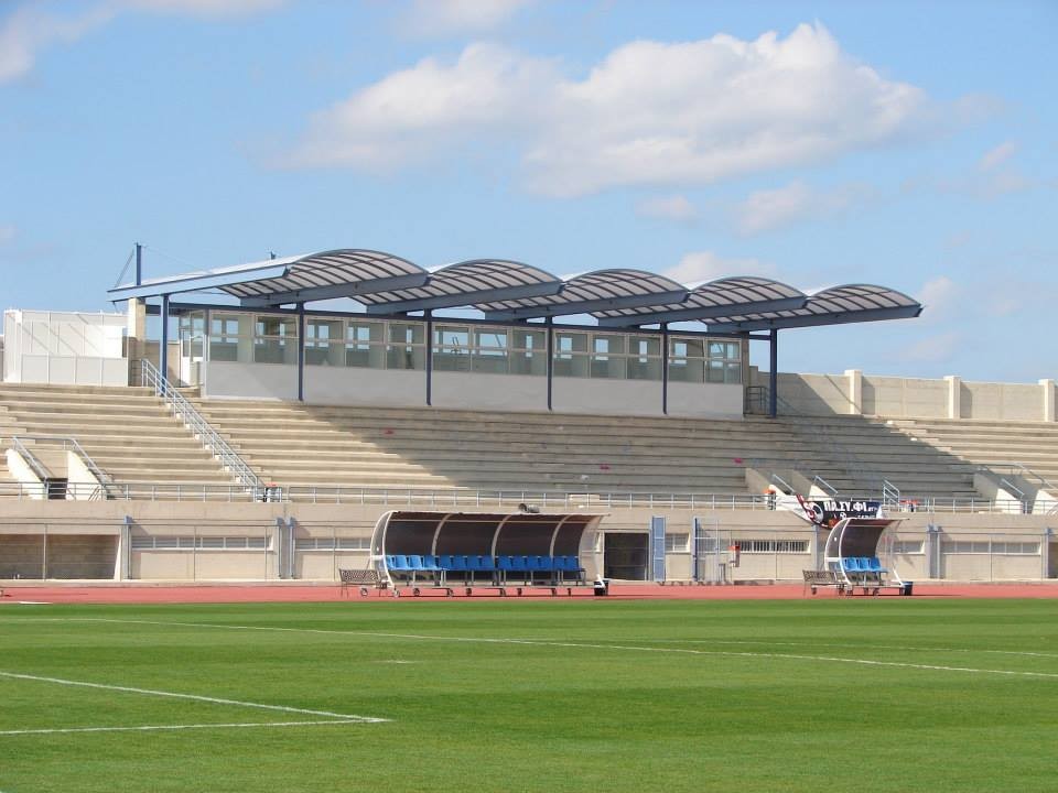 Football stadium with a grass pitch, team benches and a covered main stand.