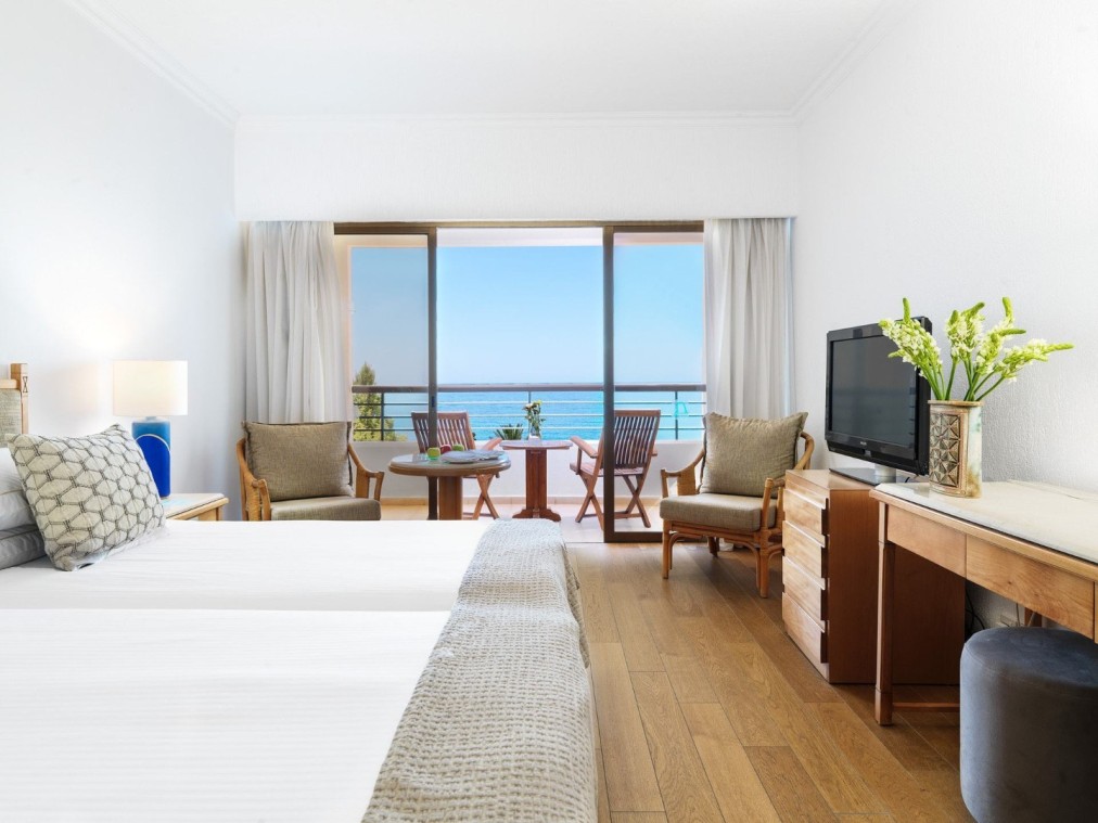 Ocean-view hotel room with balcony, wooden floor, and seating area for athletes’ rest