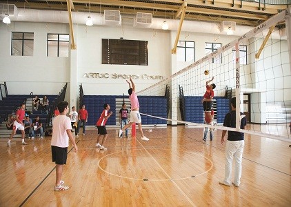 Indoor volleyball match on a wooden court, players jumping at the net and spectators on benches.