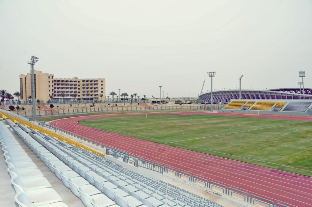 View from white bleachers of red athletics track and green infield, floodlight poles and modern stadium, training camp