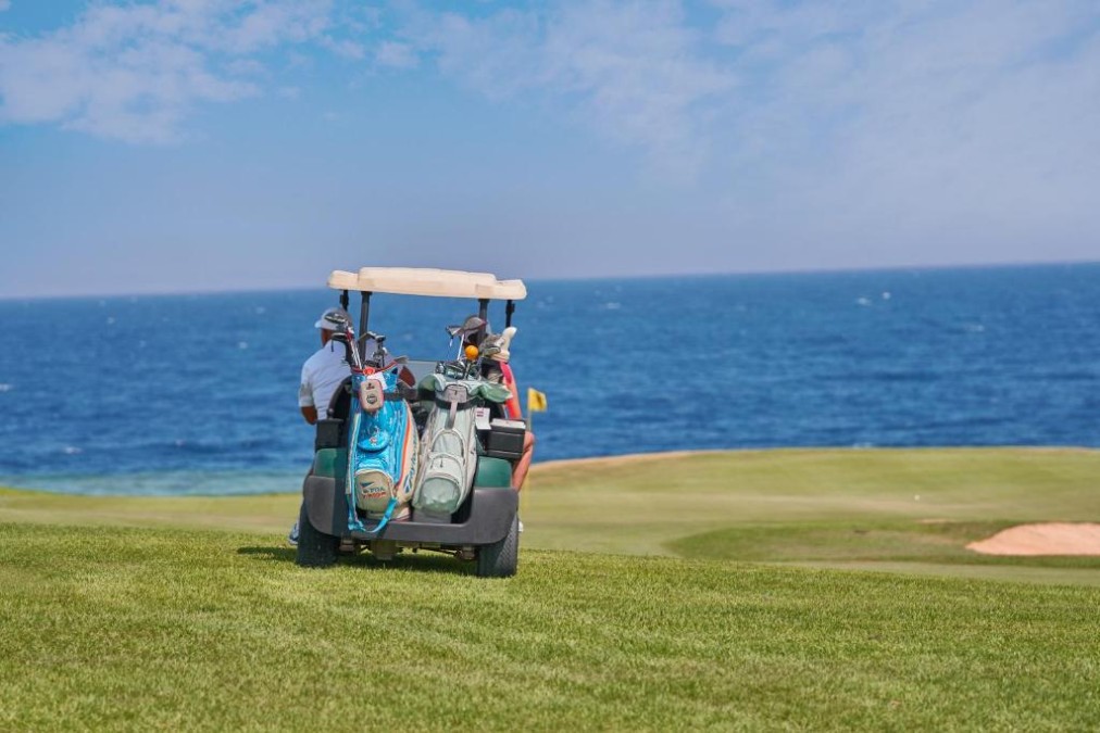 Golf cart with bags on a coastal fairway, ocean backdrop; golfers preparing for a practice round.