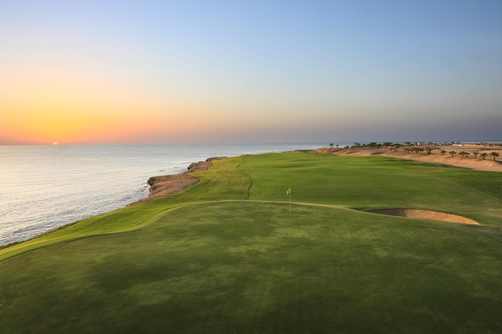 Coastal golf green at sunrise with flagstick and bunker beside the sea.