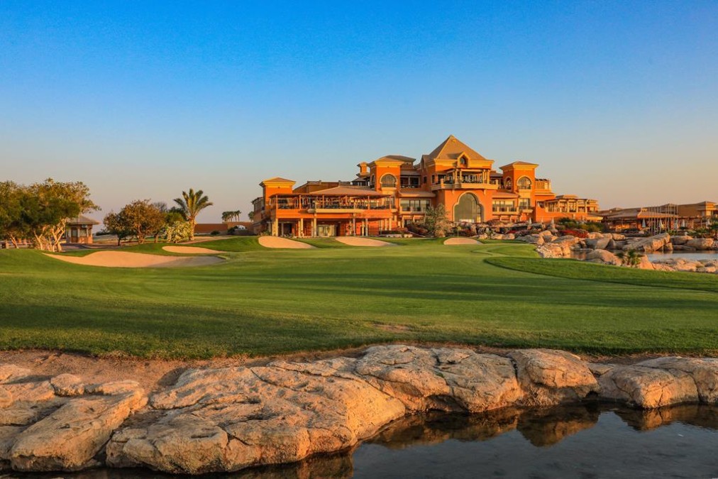 Clubhouse overlooking the fairway at sunset, with sand bunkers and a rocky pond in the foreground.