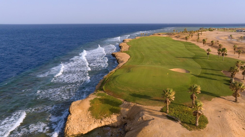 Aerial view of a clifftop green above ocean waves, with flagstick and bunker.