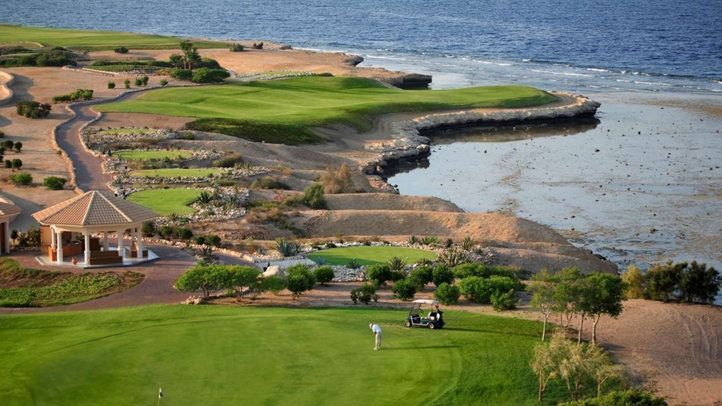 Elevated view of a coastal golf hole with gazebo, golfer, cart, and rocky shoreline.