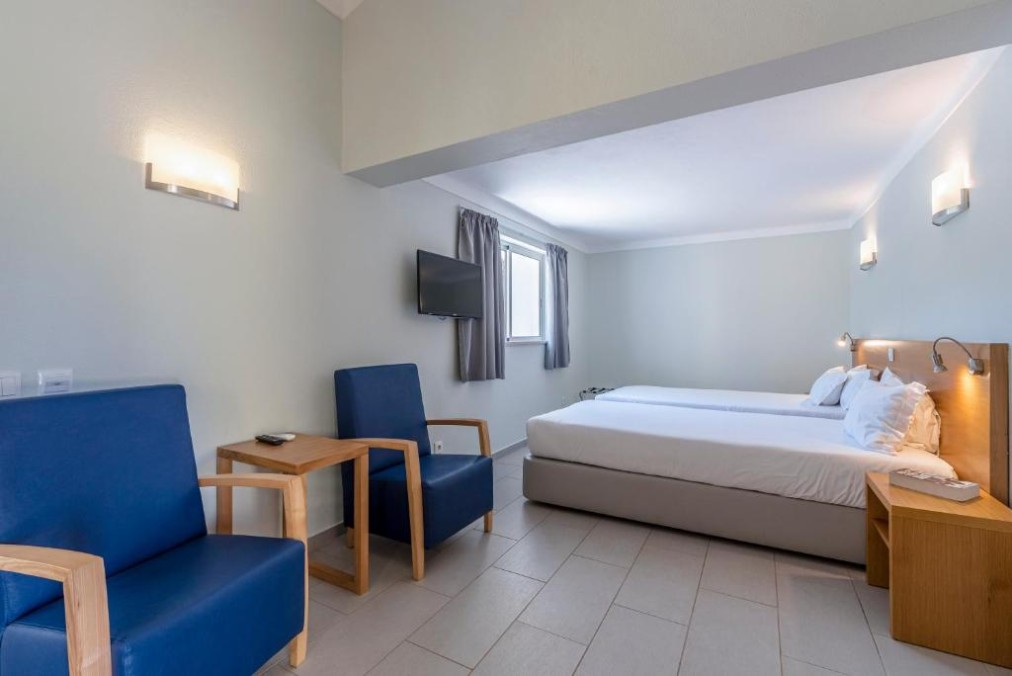 Bright twin guest room with two single beds, wall-mounted TV, and seating area with blue armchairs.