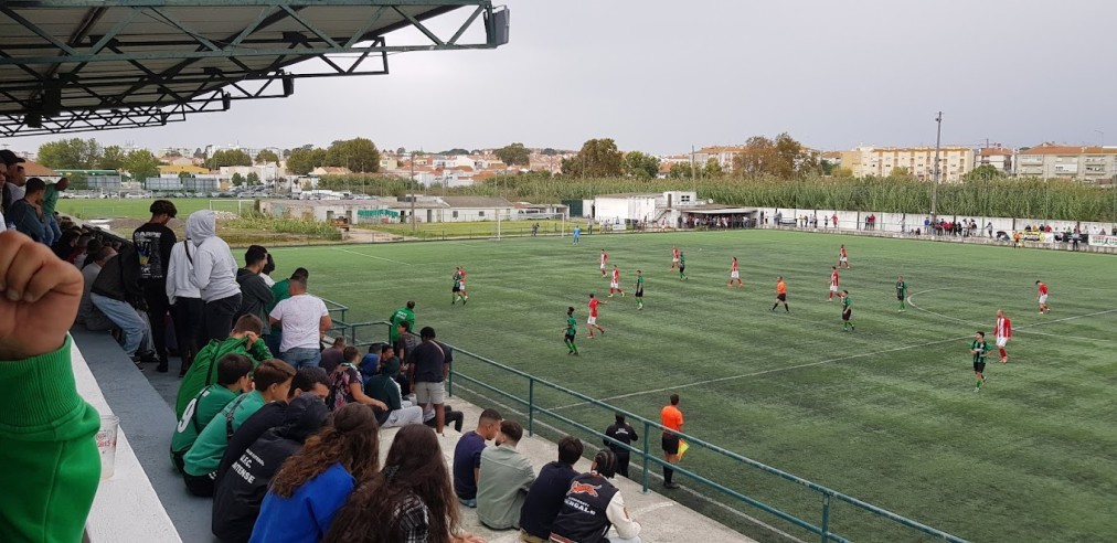 Spectators watch a football match on an artificial turf pitch from a covered stand.