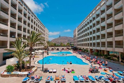 Outdoor hotel pool area with sun loungers between two large apartment-style buildings.
