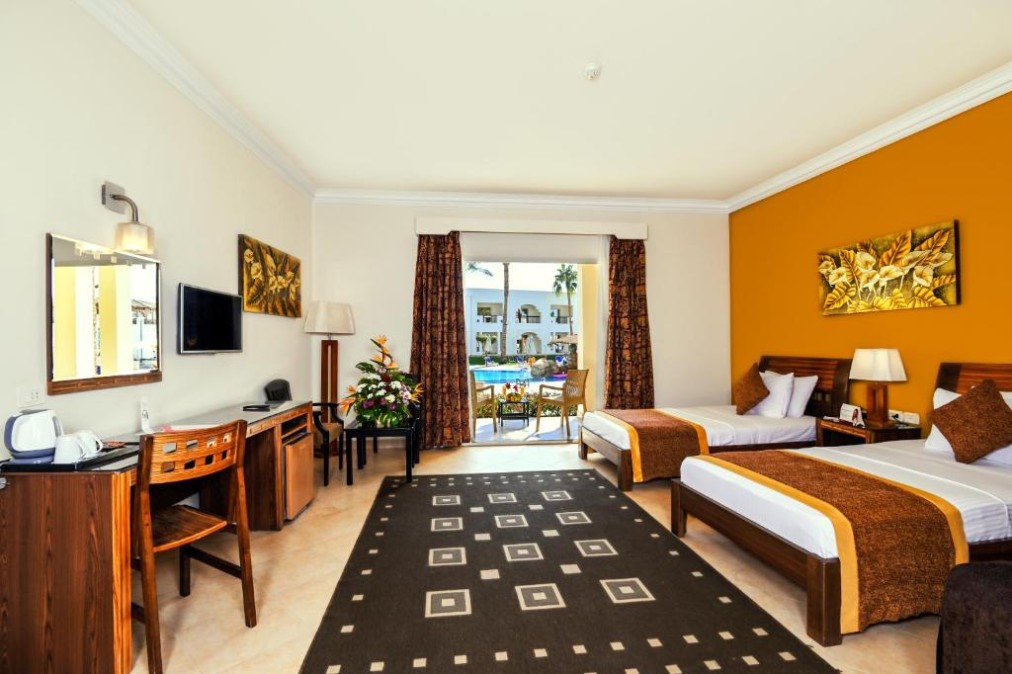 Spacious hotel room with twin beds, wooden furniture, and balcony view of pool area and palm trees