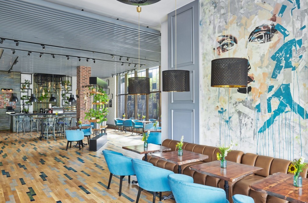 Modern hotel lounge with teal chairs, bar counter, pendant lights, and mural wall for training camp downtime