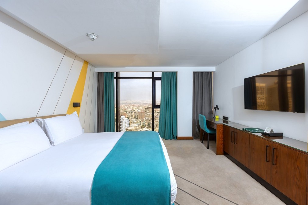 Modern hotel room with white bed and teal runner, desk and TV, large window overlooking the city