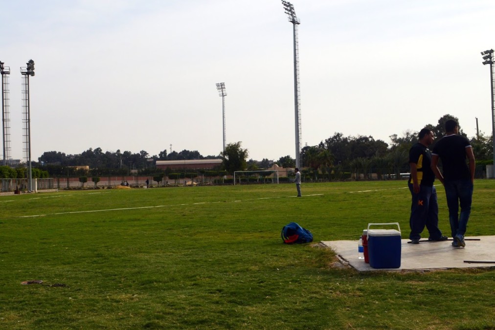 Grass football training pitch with floodlight masts; staff and equipment by the sideline.