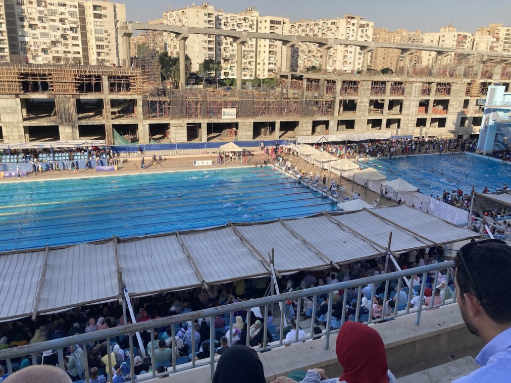 Crowded outdoor swim meet with tents along the deck and spectators in the stands.