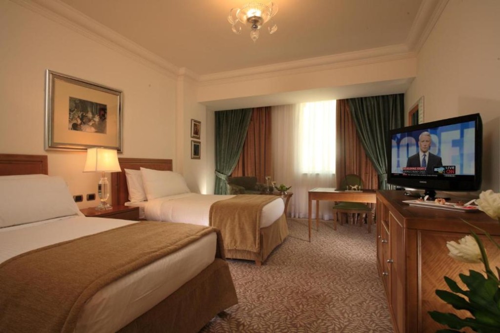 Twin room with two beds, carpeted floor, desk, and TV cabinet near a curtained window.