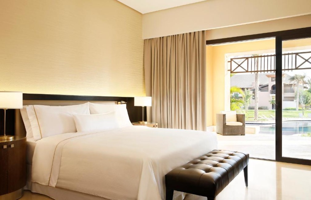 King guest room with padded headboard, bench at the foot of the bed, and terrace access facing the pool area.