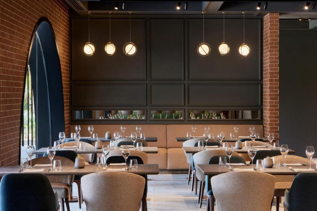 Modern restaurant dining room with set tables, brick walls, and pendant lights.
