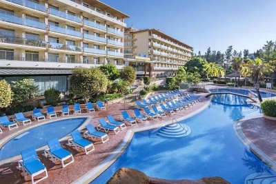Outdoor pool area with a large curved swimming pool, sun loungers, palm trees, and the hotel buildings in the background.