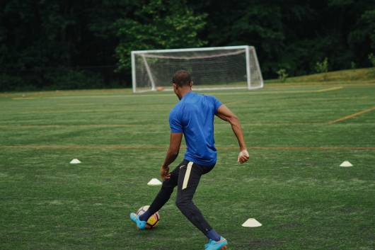 Soccer athlete in blue shirt performs dribbling drill around cones on grass field during training camp practice session