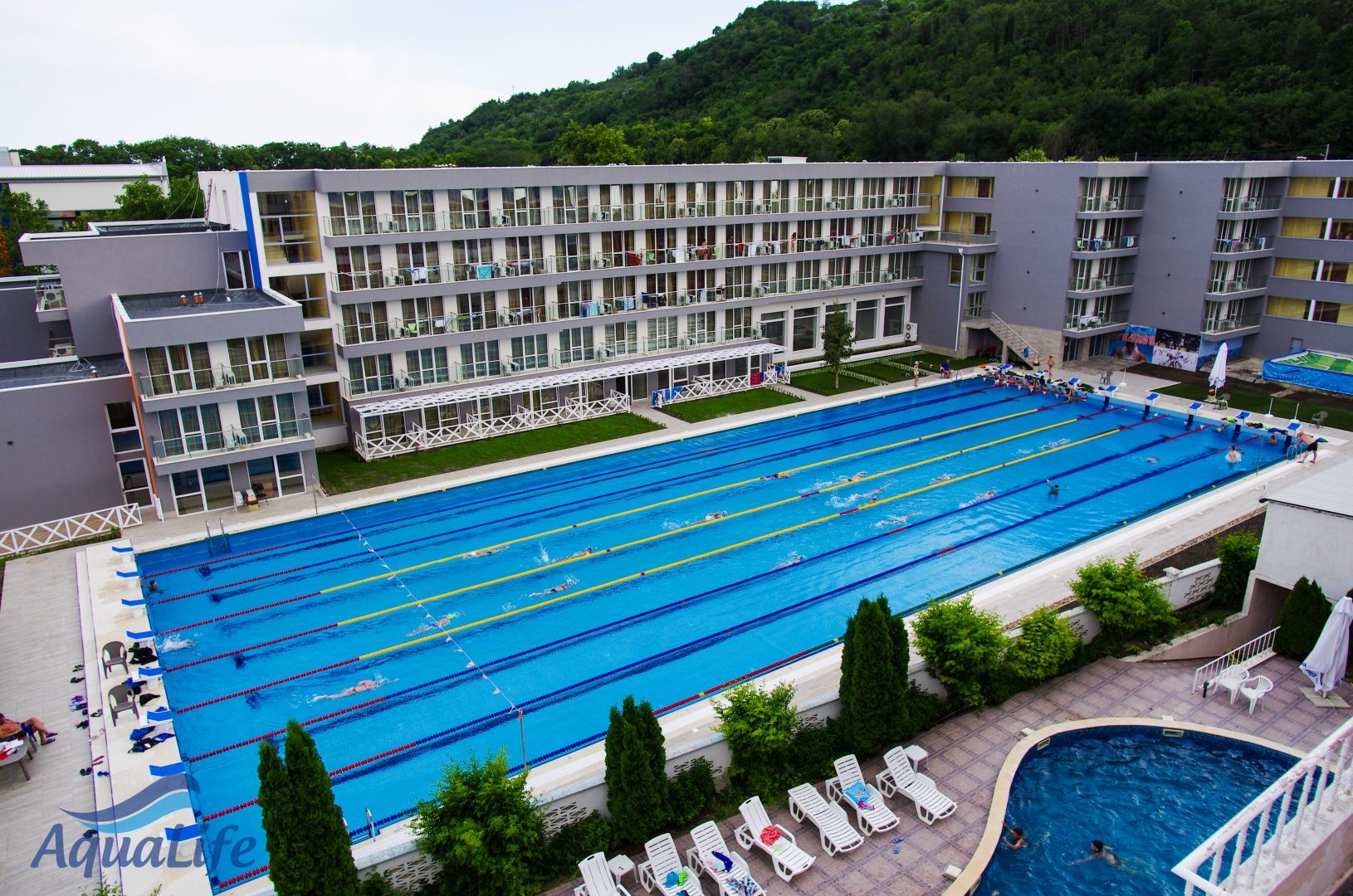 Vue en hauteur d'une longue piscine extérieure entourée d'un hôtel et de transats pour la récupération en stage d'entraînement