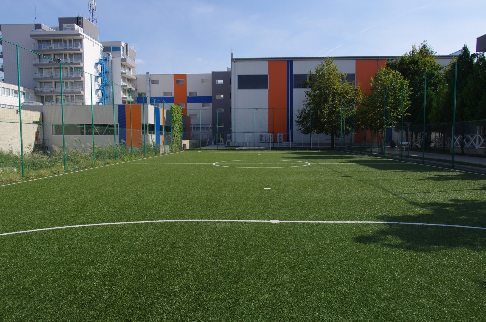 Mini-terrain de football vert avec lignes blanches, entouré de bâtiments du campus pour les exercices du stage d'entraînement