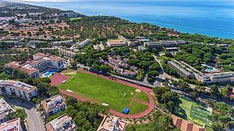 Aerial overview of a coastal sports complex with athletics track, fields, and courts.