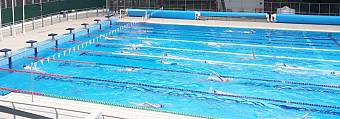 Wide shot of Olympic-size swimming pool with lanes, seating stands, and clear blue sky