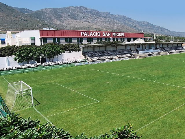 Terrain de football vu depuis une tribune couverte avec bancs le long de la ligne de touche.