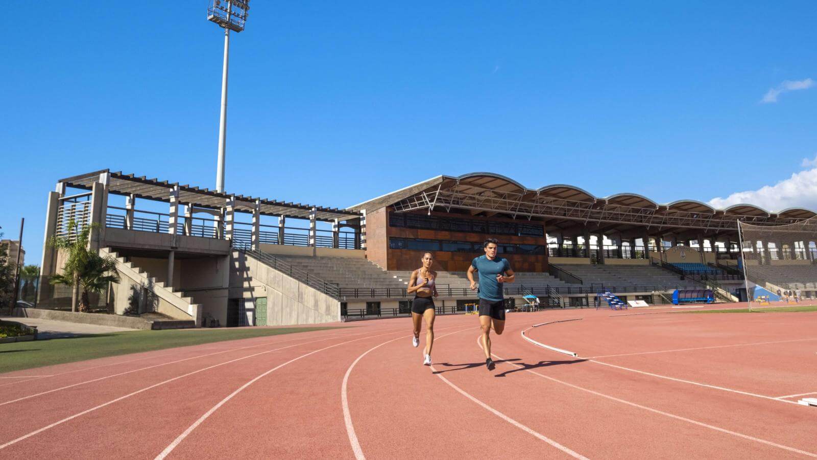 Deux athlètes courant sur une piste d'athlétisme extérieure devant les tribunes pendant l'entraînement.