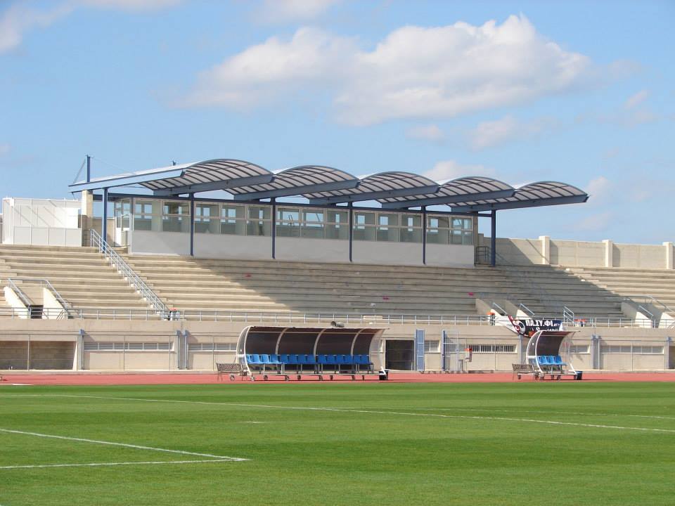 Fußballstadion mit Rasenplatz, Mannschaftsbänken und überdachter Haupttribüne.