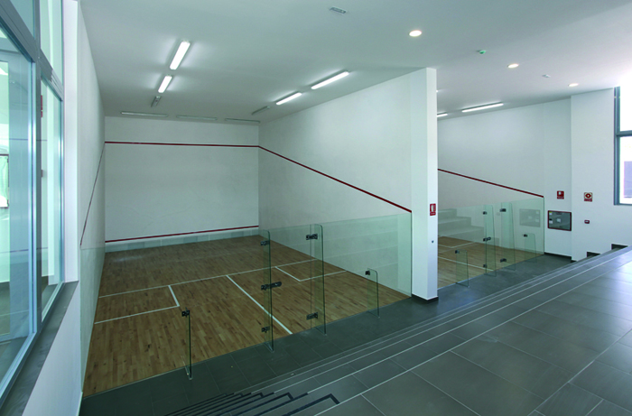 Indoor-Squashcourt mit Glasrückwand, Holzboden und Deckenbeleuchtung.
