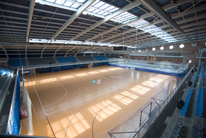 Große Indoor-Sporthalle mit markiertem Spielfeld, Tribünen und Oberlichtern.