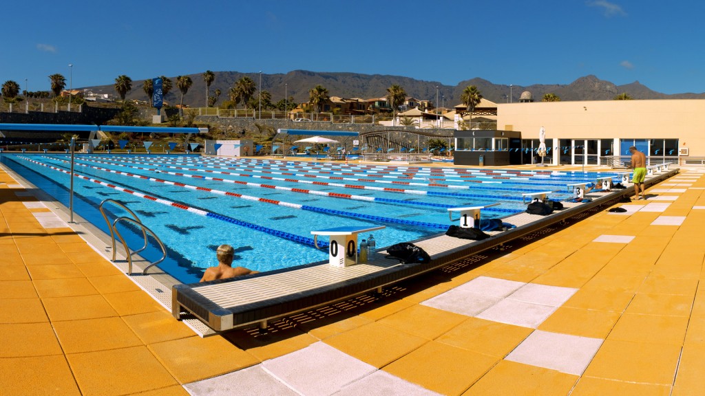 Outdoor lap pool with lane ropes and starting blocks on a wide deck, mountains in the background.