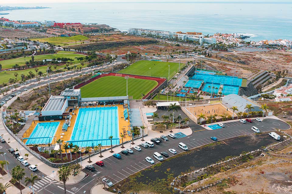 Aerial view of the coastal sports complex showing the 50 m outdoor pool, football pitch with running track, multiple racket courts, and adjacent parking area.