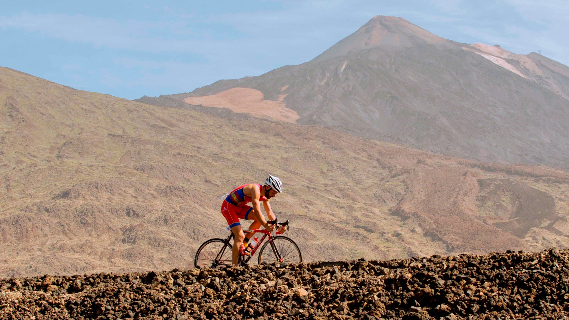 Road cyclist riding on volcanic terrain with a mountain backdrop.