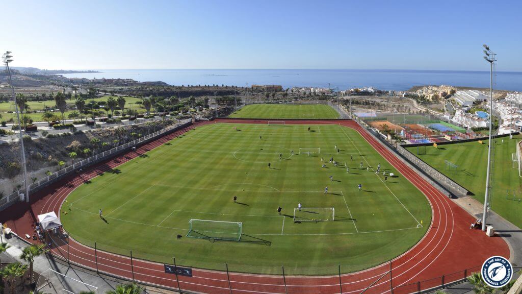 Football training session on a full-size pitch inside a running track, ocean beyond.