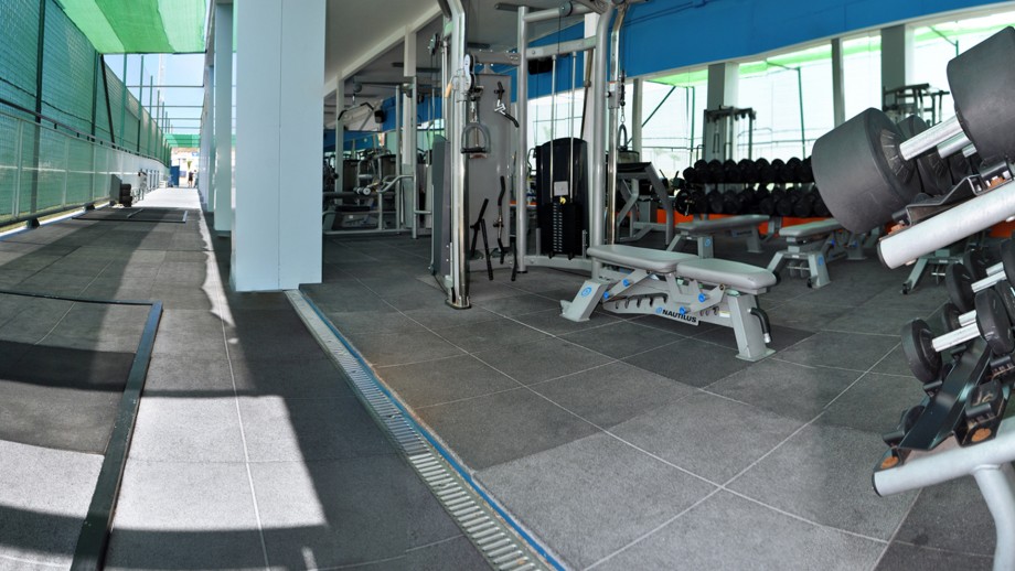 Semi-outdoor gym corridor with strength machines, benches and dumbbell racks.