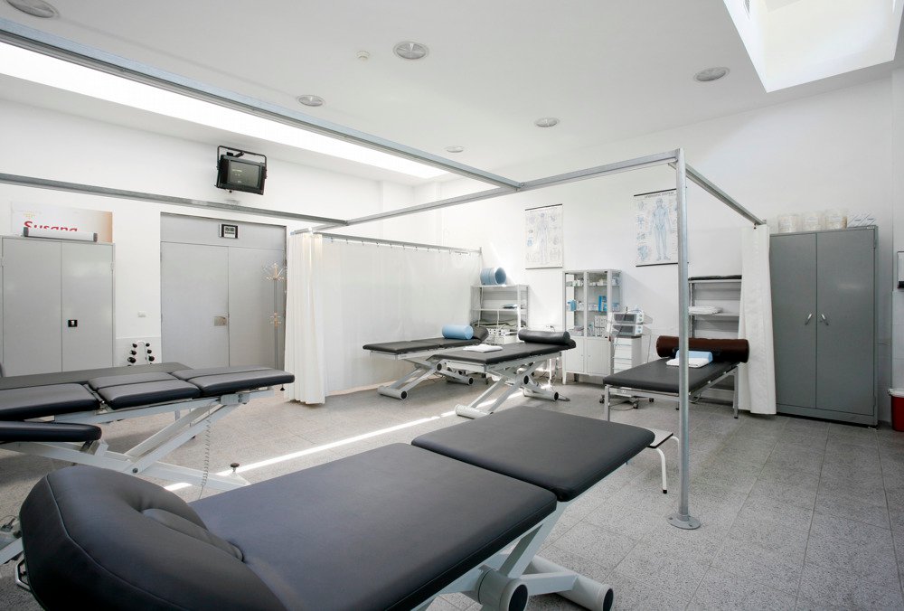 Bright physiotherapy treatment room with multiple therapy tables, privacy curtains and medical storage cabinets.