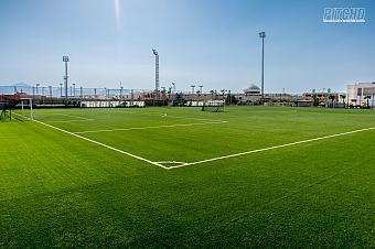 Wide view of green football pitch at training camp with tall floodlights and clear blue sky