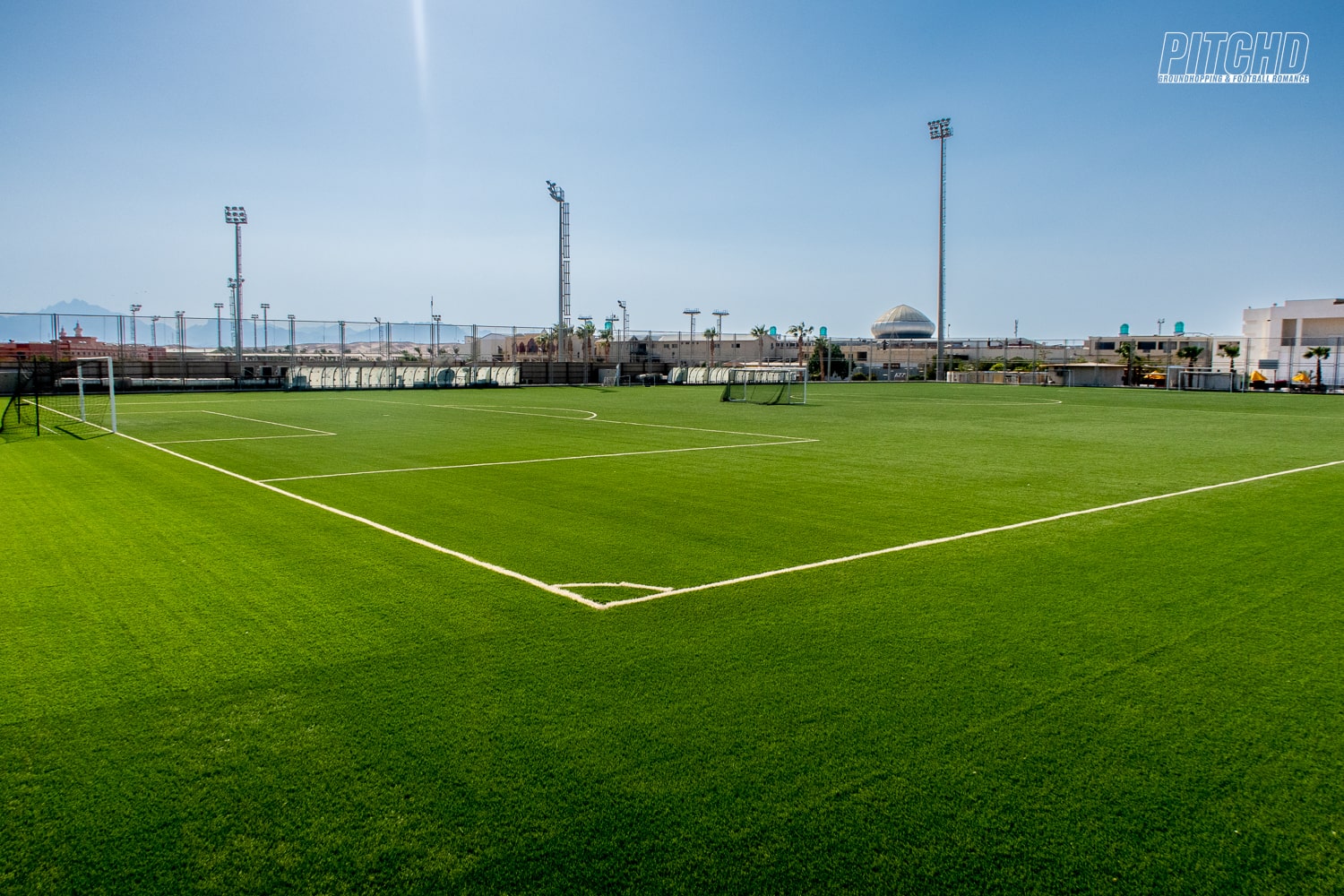 Wide view of green football pitch at training camp with tall floodlights and clear blue sky
