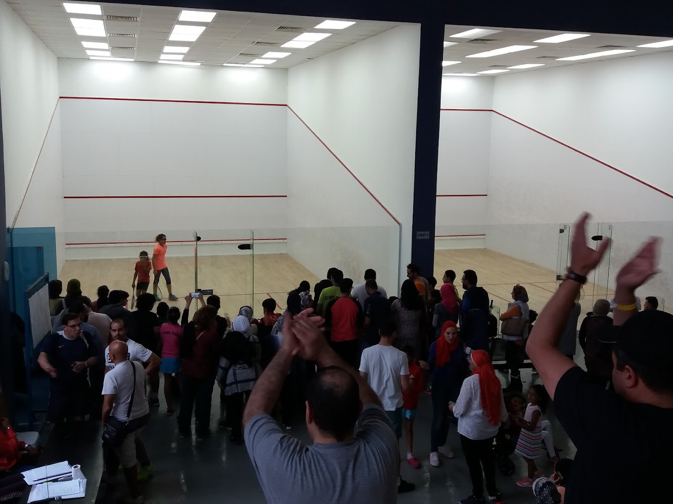 Spectators watching a squash match on an indoor glass court, applause during play.