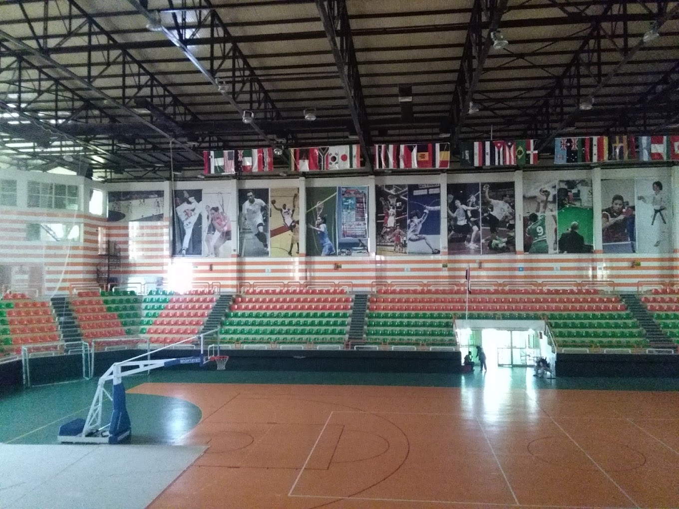 Panoramic indoor sports hall with court, basketball hoop, spectator seating, and national flags along the rafters.