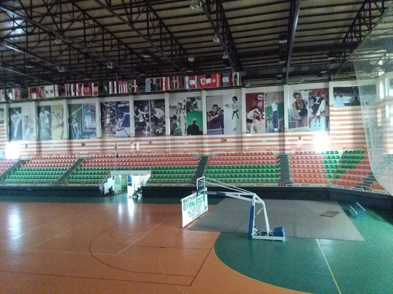 Wide view of an indoor arena showing court markings, basketball hoop, tiered seating, and sports banners.