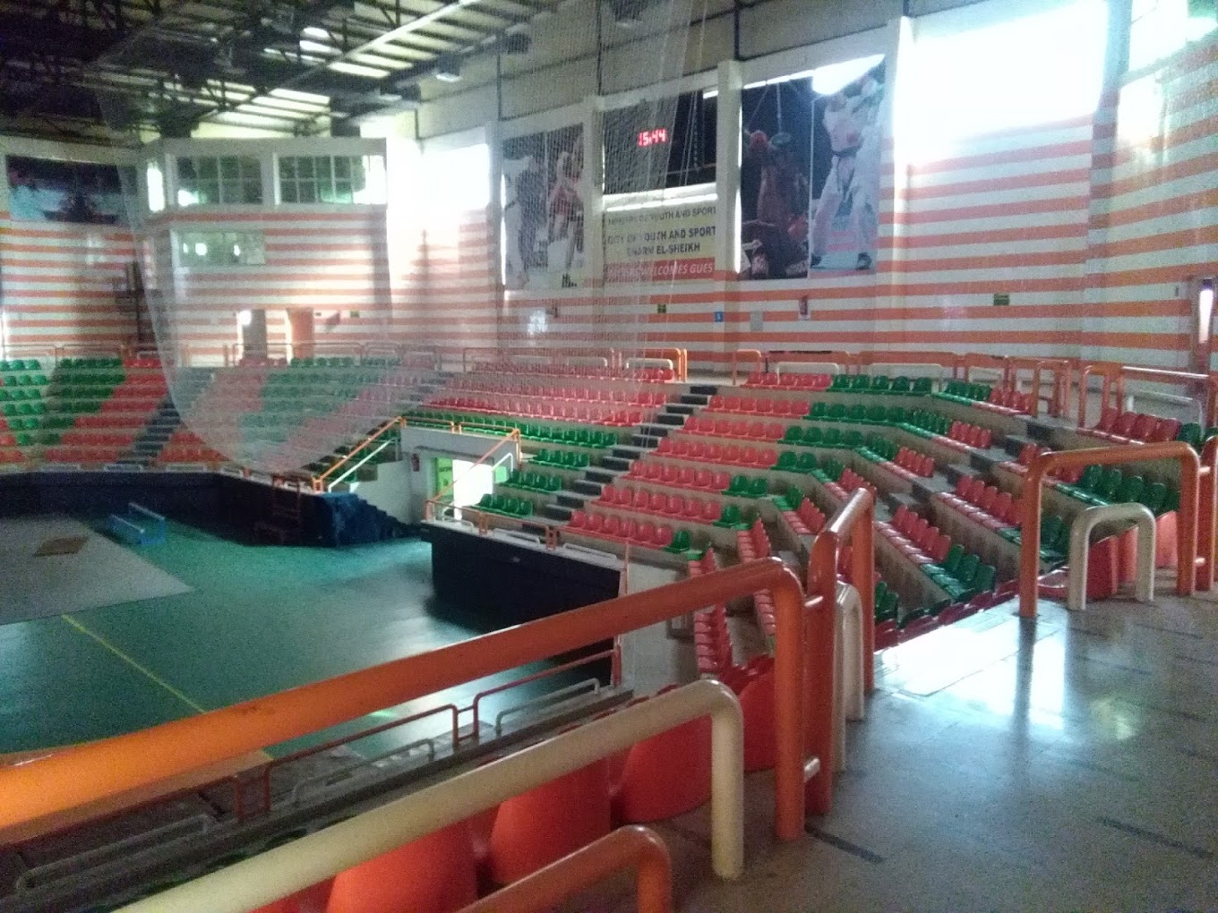 Upper-tier view of an indoor sports arena with red-green bleachers, wall murals, and a digital scoreboard.