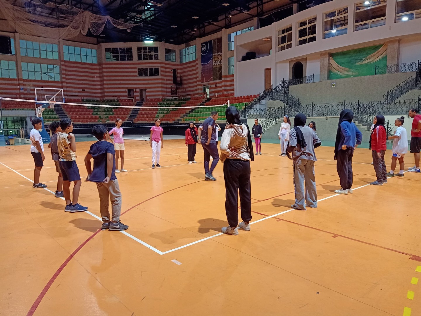Athletes in a circle on an indoor court during volleyball training camp warm-up; tiered stands in background.
