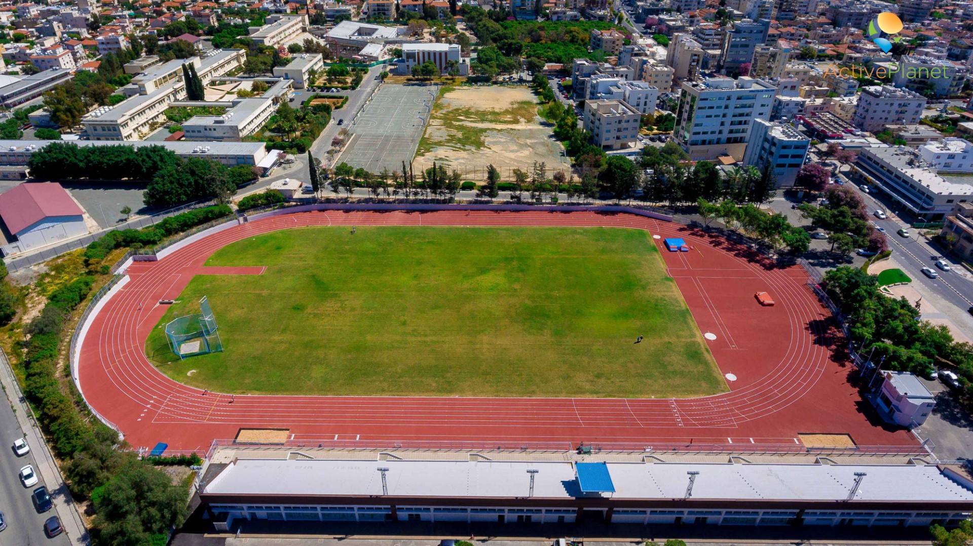 Aerial view of a red athletics track and green field surrounded by city buildings, used for training camps and practice sessions