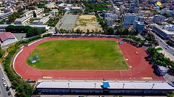 Aerial view of a red athletics track and green field surrounded by city buildings, used for training camps and practice sessions
