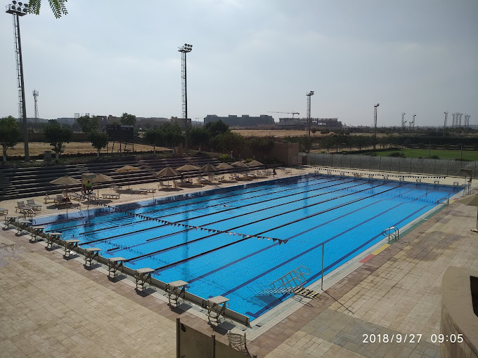 Wettkampf-Freibad mit Bahnleinen und Startblöcken, umgeben von Sitzreihen und Sonnenschirmen.